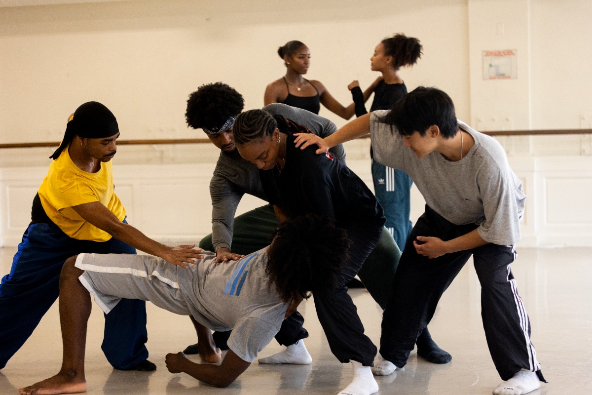 A group of dancers in a brightly lit studio are captured during rehearsal. In the foreground, a barefoot male dancer in a gray t-shirt and gray shorts is parallel to the floor and facing up, using his feet and one arm to hold himself up in a pose reminiscent of a crab walk. Four other males dancers in casual practice clothes are grouped around him, providing physical support or holding their hands over his torso. In the background, two female dancers in casual practice clothes are interacting near a studio wall.