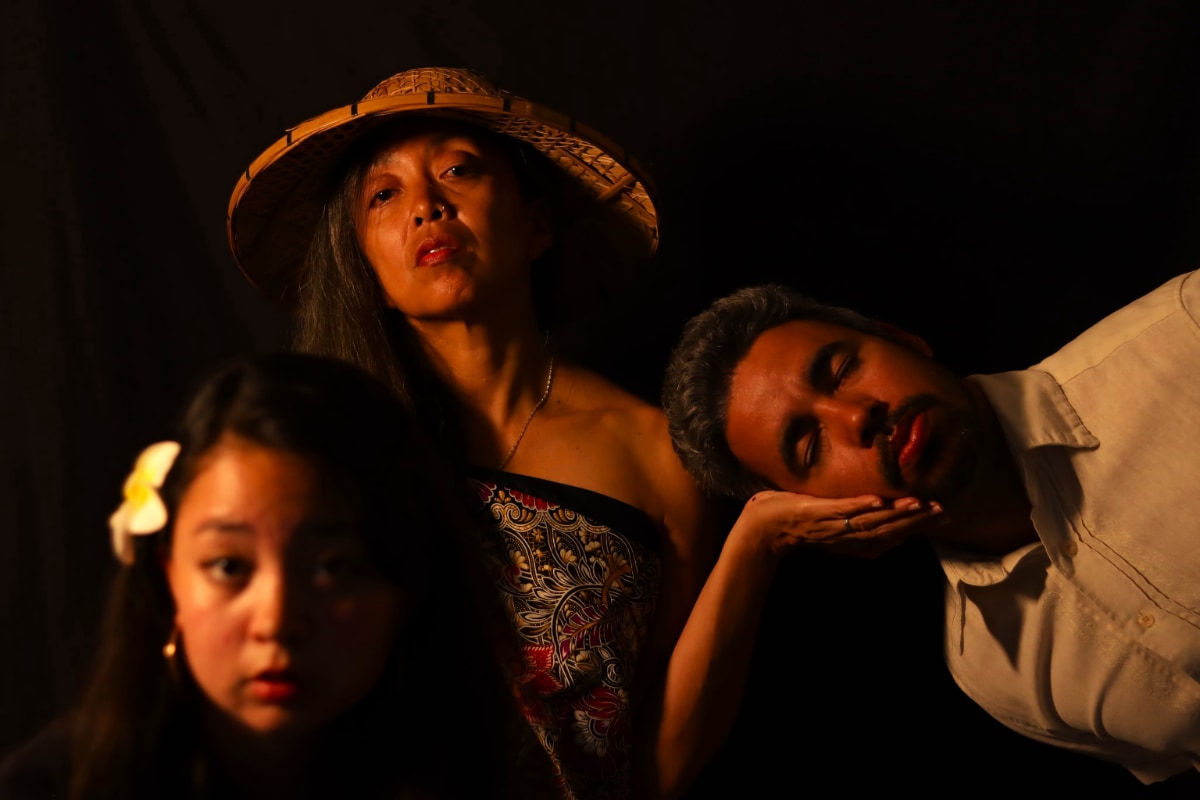 A dramatic, low key studio portrait features three people set against a solid black background. The scene is illuminated by a warm, golden-hued light source from the left side, which casts deep shadows and emphasizes the rich skin tones and textures. In the lower left foreground, slightly out of focus, a young woman with a white flower in her dark hair looks toward the viewer. Behind her in the center left, in focus, a woman wearing a conical straw hat and a patterned wrap top gazes forward, her head tilted back slightly. She holds one hand with the open palm facing up. A man in a plain, light colored button-down shirt leans in from the right and rests his head, eyes closed, on her open hand.
