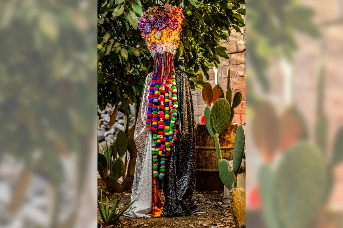 A person entirely hidden in a costume stands outdoors in front of a leafy green tree, several prickly pear cacti, a weathered metal barrel, and a brown brick wall. The figure is draped in a long cloak made of two complementary fabrics: the left side is white with black spots similar to alligator print, and the right side is the same but with the colors inveterd. Near the ground and barely visible between the two panels is a glimps of rich orange satin. Half a dozen long strands of plum-sized, multi-colored pom-poms drape down the front and hang past the knees. The person is wearing a two-foot tall, inverted conical headress that is wider at the top. It's covered with large flowers that are cream colored near the person's head, then bright yellow in the middle, and alternating shades of purple and red around the top half. Thick braid-like strands of fabric or yarn in various colors hang from the brim of the hat like a veil, completely obscuring the person's face. 