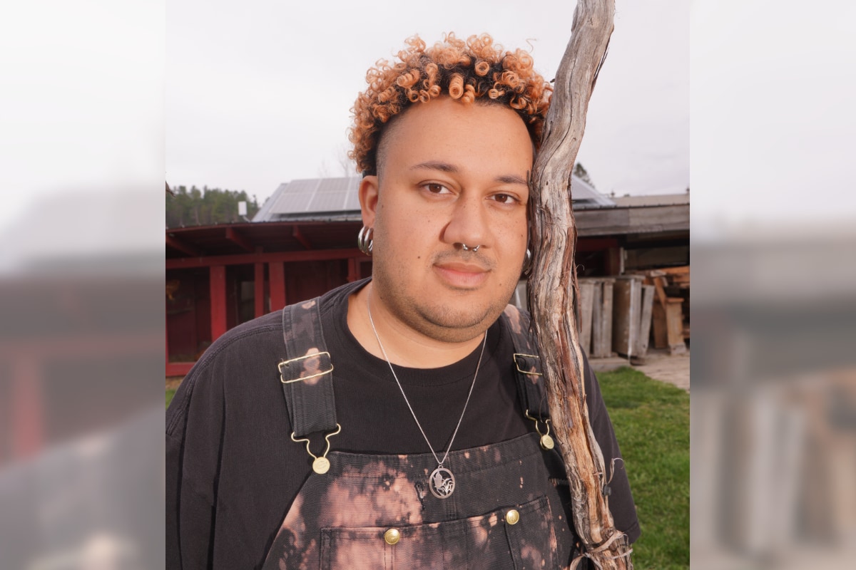 A young person with slight beard stubble stands outside on an overcast day and smiles gently at the viewer, with their forehead pressed gently against a weathered, dried-out tree branch outdoors that they appear to be holding like a staff or tall walking stick. The person has short, curly, copper-colored hair with black roots, wears silver hoop earrings and a silver circular barbell septum piercing, and is dressed in black-and-white tie-dyed overalls over a black t-shirt, accented with a silver pendant necklace. The background shows a rustic setting with a red structure, solar panels, and a grassy lawn, indicating a farm or rural location.