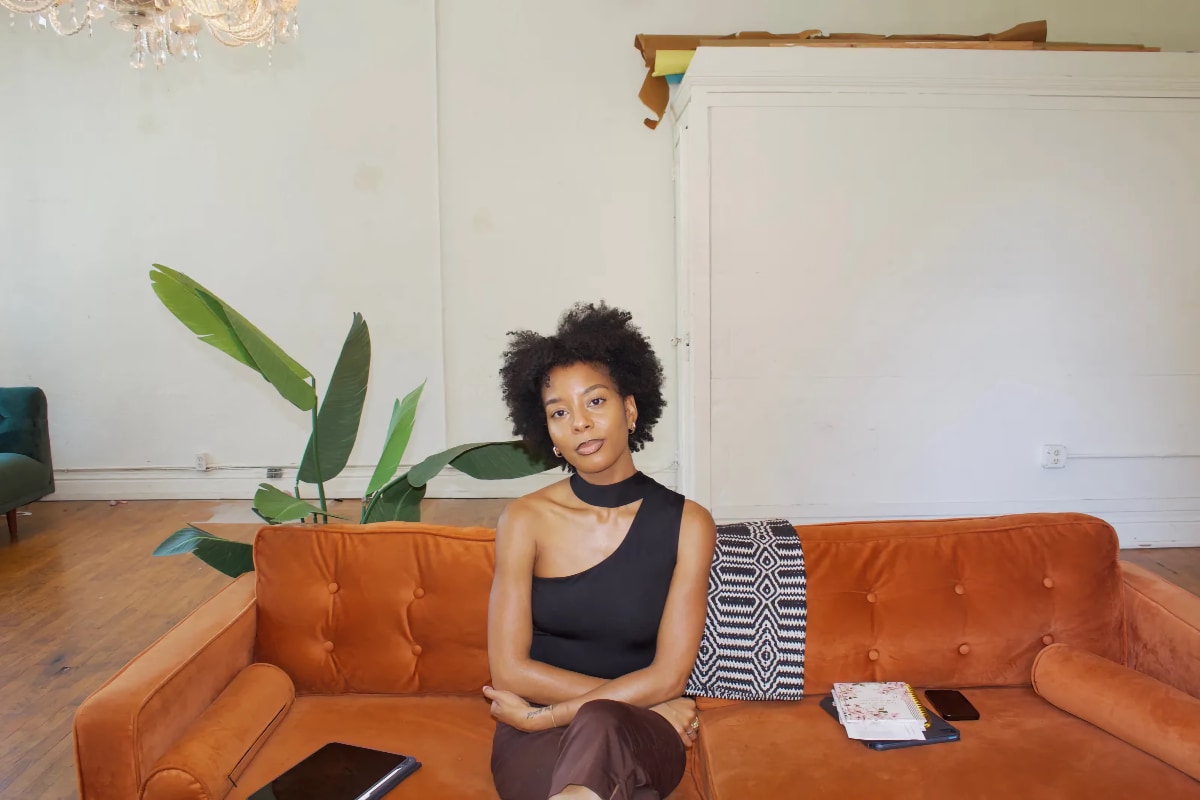 A Black woman with natural mid-length hair in a center part, and wearing a black one-shoulder top with a neck strap and brown capri pants, sits composed on a tufted, burnt-orange velvet or suede sofa with her arms folded across her waist and her legs crossed and stares serenely at the viewer. An iPad with the screen off is on the sofa to the left of her, and a paper notebook and phone are to the right of her. The location appears to be a bright, sparsely decorated apartment or studio, featuring white walls, a large potted plant behind the couch, and the edge of a green chair in the background on the left.