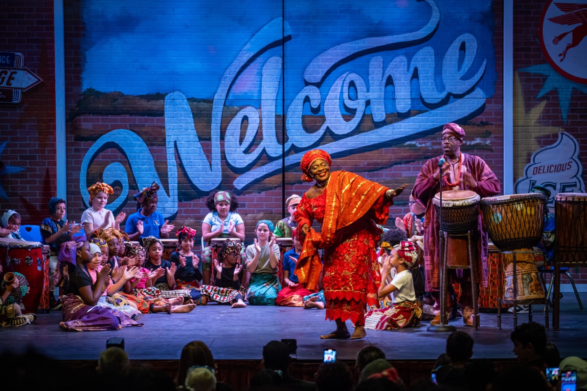 A photograph captured from the audience's perspective of a lively stage performance. The backdrop is a painted brick wall featuring a large, blue script sign that says "Welcome." In the center of the stage, a brown-skinned woman in African clothing dances with arms outstretched. To her right, a brown-skinned man in African clothing accompanies her with a drum. Seated on the floor in a semi-circle behind them is a large group of children dressed in bright, multicolored African clothing, chanting and clapping their hands in response to the drum call.