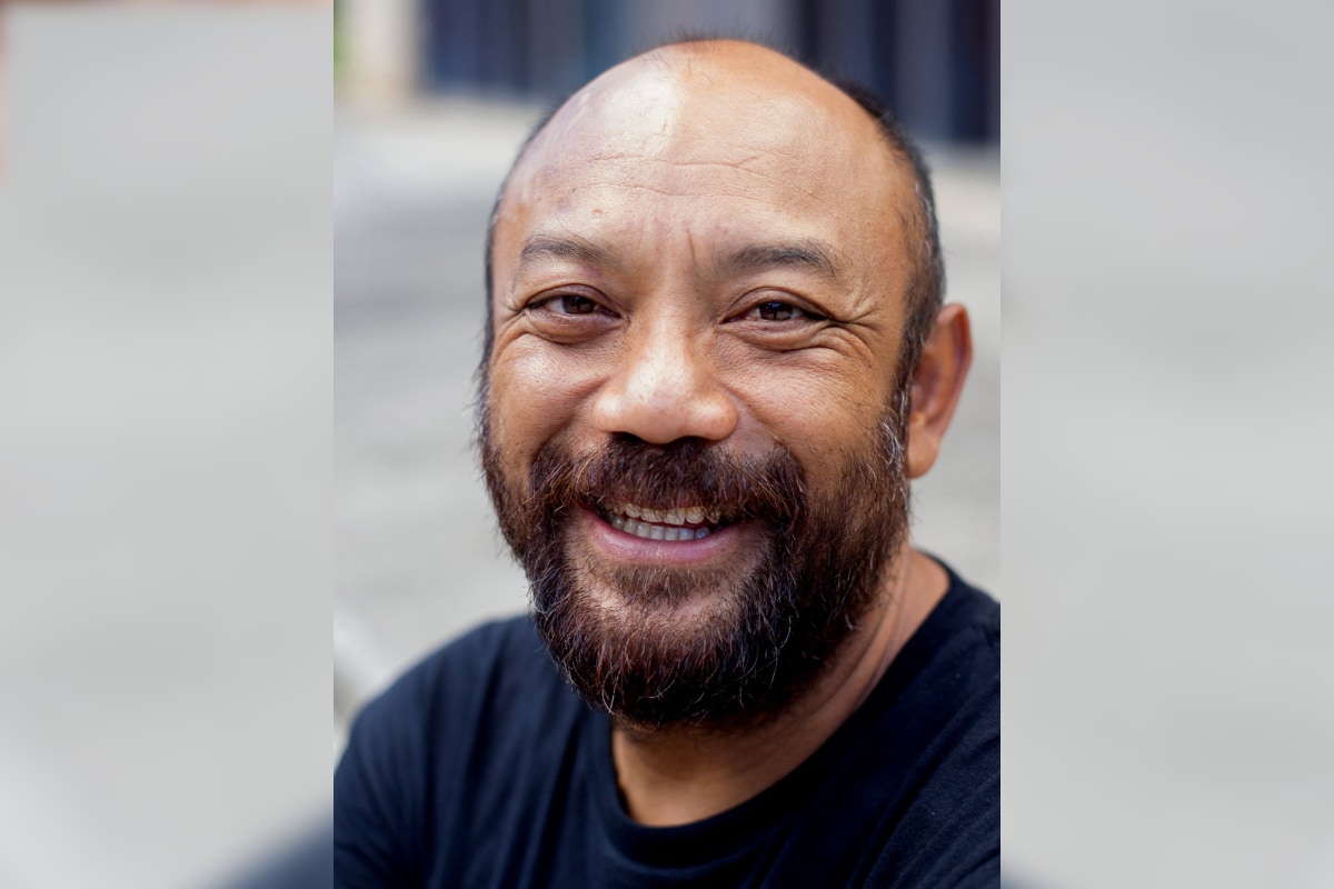 A close-up portrait of a man with a warm, smiling expression. The background is soft and out of focus, and suggests a sidewalk or plaza in a city. The subject has a short, dark beard, expressive brown eyes, and is wearing a simple black t-shirt.