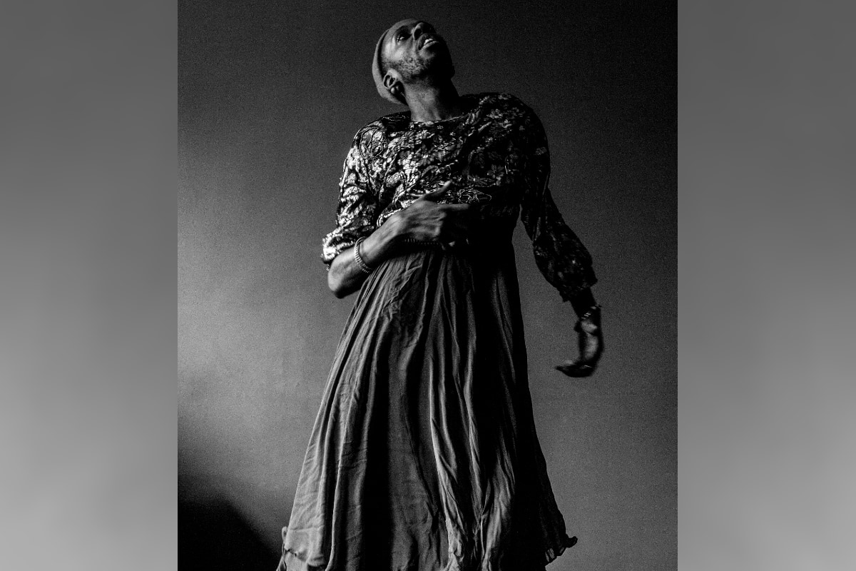 A dramatic, high-contrast black-and-white studio photograph shows the performer Raja Feather Kelly in a long, full-skirted dress with a textured or patterned bodice and a beanie. They are looking upward with an open, emotive expression, with one hand across their torso and the other extended down and back. The background is a plain, dark studio backdrop.