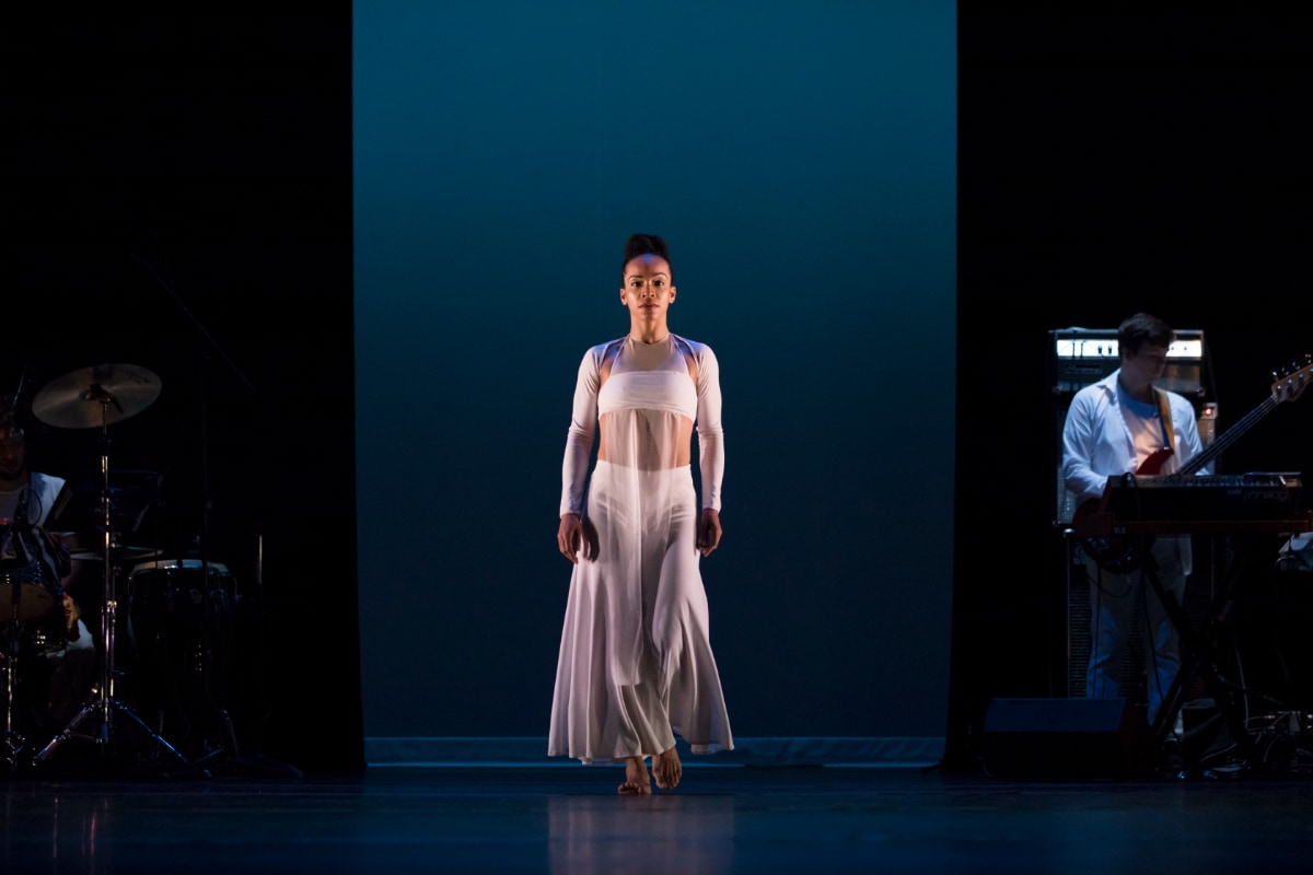 A photograph of a stage performance shows a female, medium skinned dancer centered under cool blue lighting. She is wearing a flowing, sheer white costume, and appears to be slowly walking toward the audience. The wall behind her is a cool teal-blue. On the left, in near darkness, a drummer is partially visible, and on the right, a musician plays an electric bass guitar with a keyboard nearby.