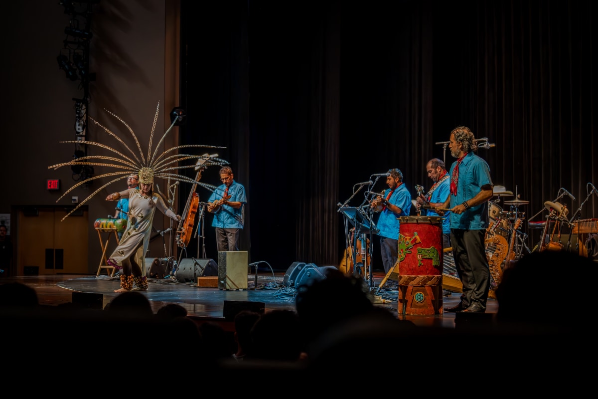 A photograph taken from the audience's perspective shows a dancer and five accompanying musicians on a warmly lit theater stage. On the left, a medium-skinned Latino dancer in a white traditional Mexica, or Aztec, dance garment performs with her arms outstretched, wearing an elaborate feathered headdress with long golden and dark plumes fanning dramatically upward and outward. In her right hand she holds a Mexica, or Aztec, traditional rattle, called ayacaxtli in Nahuatl. She has seed rattles, or ayoyotes, around her ankles. Behind and to the right and left, the five Latino musicians, wearing dark pants, turquoise blue shirts, and red bandanas around their necks, stand and play traditional Mexican or Mesoamerican instruments, including — in the foreground on the right side of the image — a large, vividly painted red ceremonial drum decorated with animal figures in green and gold. 