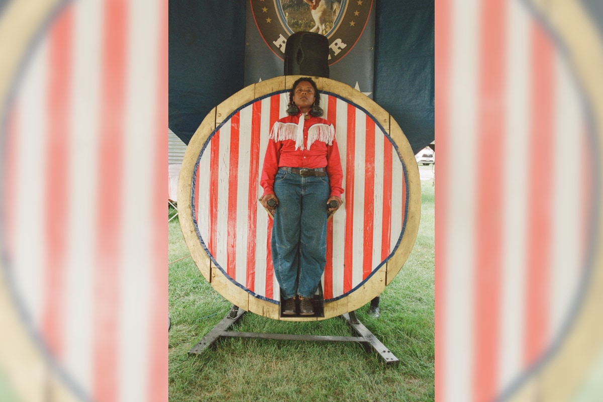 A portrait of a dark-skinned woman standing on a small platform attached to a large, circular wooden knife-throwing target painted with red and white stripes. The woman has shoulder-length black hair and is dressed in rodeo attire, including cowboy boots, blue jeans, a brown leather belt, a red long-sleeve shirt with white fringe across the chest, and a white bandana tied around her neck. Her expression is calm and focused, and she holds firmly onto handles on either side of her hips as if preparing for the wheel to spin. The location is a grassy area underneath a blue tarp at what looks like a fair or festival.