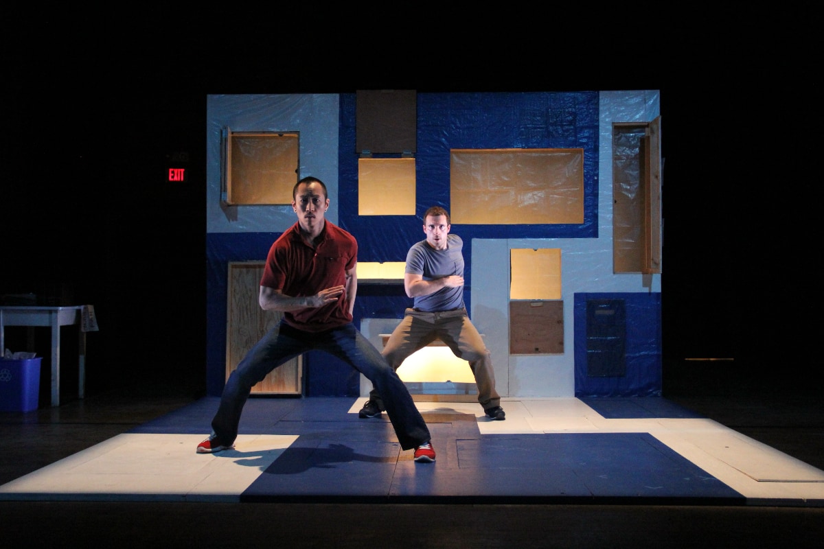 Two male dancers perform on a small stage that's painted blue but features large, irregularly positioned rectangular areas of white. The backdrop is a large panel featuring competing rectangular areas of dark blue and light blue, with several window-like openings at different heights that reveal a wall bathed in golden light. The man on the left, who is closest to the viewer, stands slightly crouched with his legs spread wider than shoulder-width apart, one arm bent in front of his torso and the other arm hidden behind his back. He is wearing a maroon polo shirt, dark blue jeans or pants, and red tennis shoes with white laces. Behind him and to the right, the other man echoes the first dancer with a matching pose. He is wearing brown pants and a blue t-shirt.