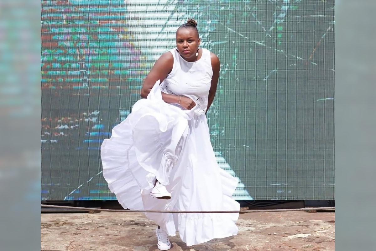 A brown-skinned woman performs a dance movement on a floating barge on a brightly lit day. She is lifting one knee while holding the flowing layers of a long white dress. She wears white low-top sneakers and hoop earrings, and her hair is pulled into a high bun. Behind her is a large digital screen with abstract lines and patterns and an overall greenish-blue cast.