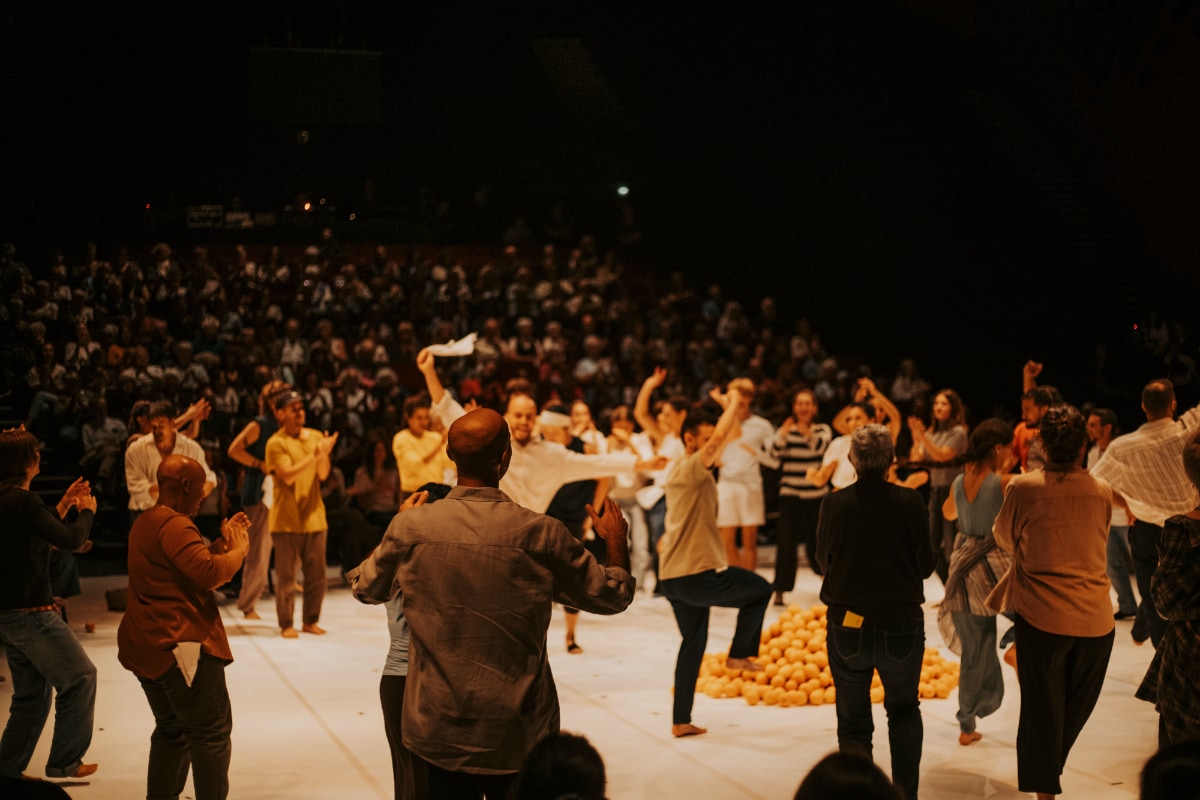 A photograph taken from the stage shows a large group of performers and audience members gathering on a white marley stage in a circle around a pile of oranges. Some are clapping, while others dance with arms raised. One performer waves a white handkerchief in the air. In the background a large seated audience is visible.