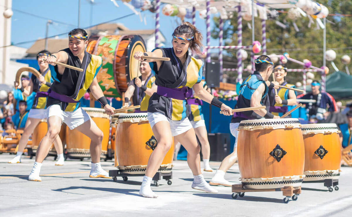 A group of young Asian-Americans of various genders perform for a crowd on a bright sunny day. They are dressed in stylized cultural costumes of black and yellow with large purple belts, white shorts, and white ankle boots. They are holding large drum sticks and striking large traditional drums while smiling at the audience out of frame.