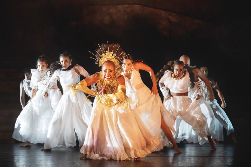 8 dancers on stage wearing white costumes with determined warrior-like facial expressions. They are in a triangle formation with the front center dancer wearing gold adornments including a large gold crown. She is rubbing her hands together and fiercely clenching her teeth.