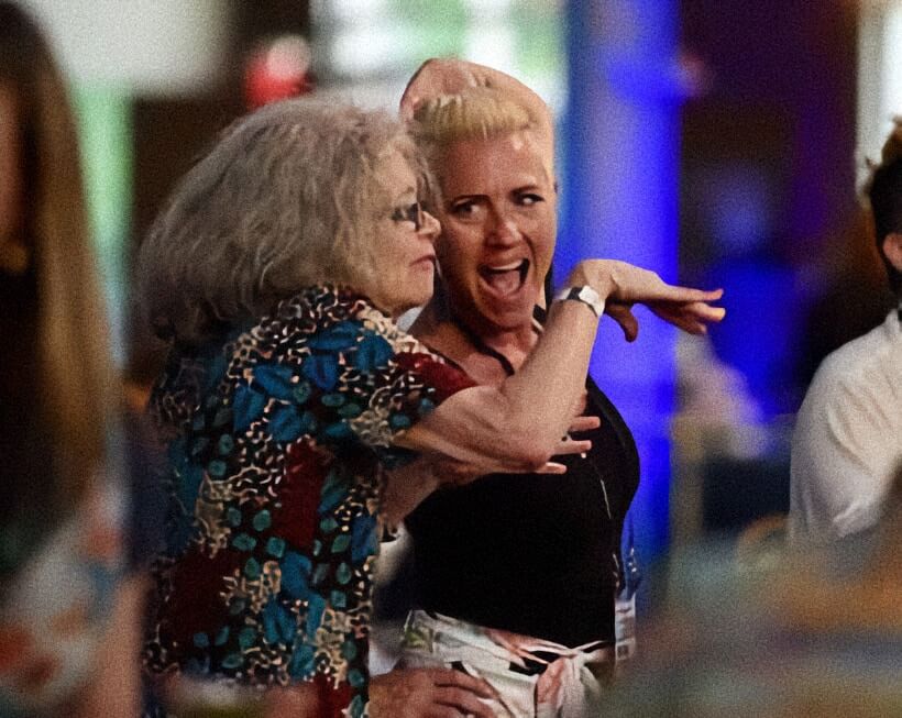 An older woman with gray hair and glasses gestures animatedly with her arm while standing beside a smiling blonde woman at an indoor event. The blonde woman reacts with an open mouthed expression as colorful lights and other attendees blur in the background. The candid moment captures lively conversation and energy during a social gathering.