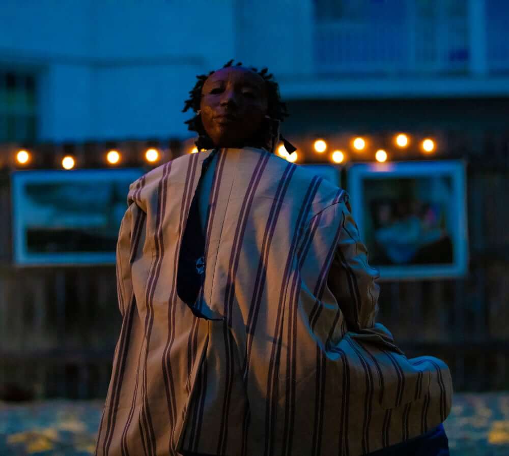 An image of a medium-skinned woman sitting down with her back to the viewer. She is wearing a mask tied to the back of her head and a tan blazer with purple stripes; her hairstyle is short locs.