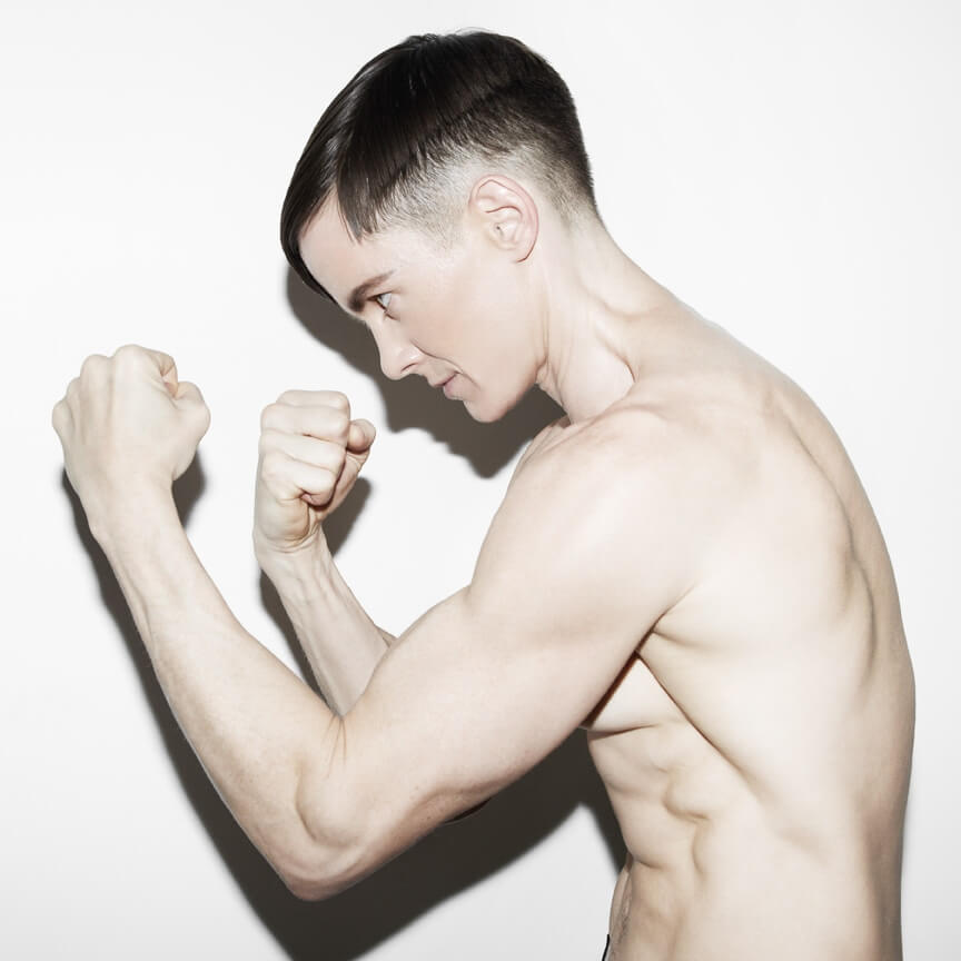 A light-skinned trans masc individual stands in profile with both fists raised as if in a defensive fighting position.