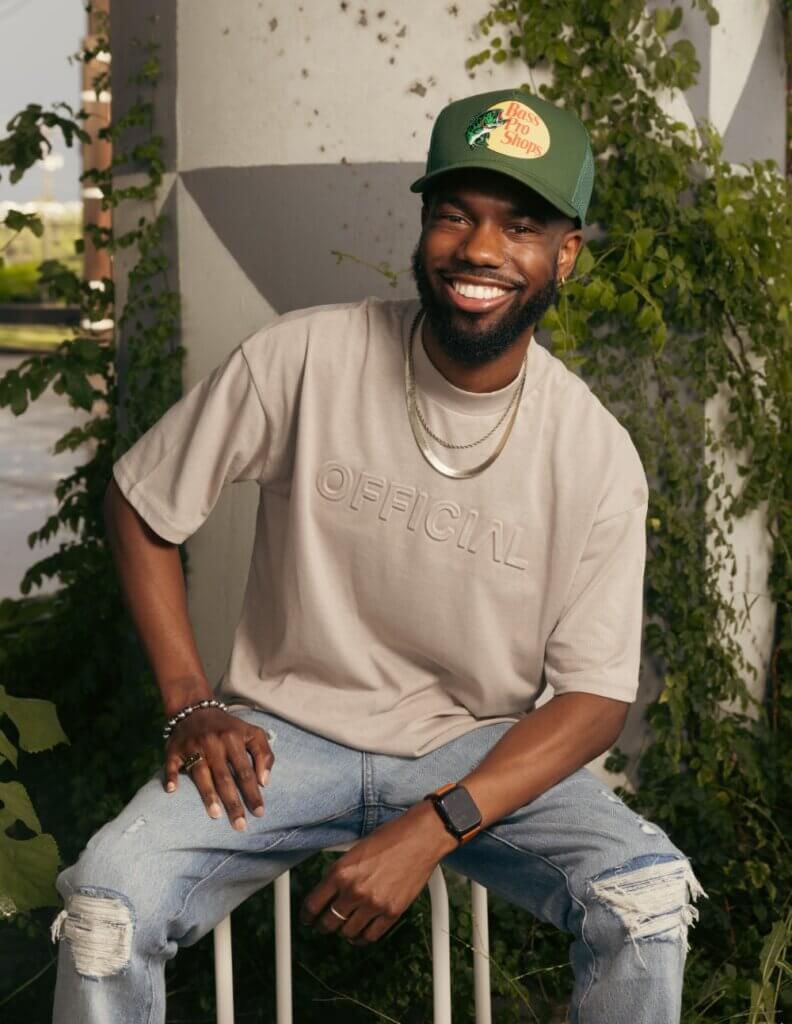 This is an image of a dark-skinned African American man with a full beard. He wears an olive-green baseball cap with a Bass Pro Shop logo, a tan shirt, blue jeans, and fashionable jewelry. He sits on a stool and looks at the camera, smiling broadly at the viewer.