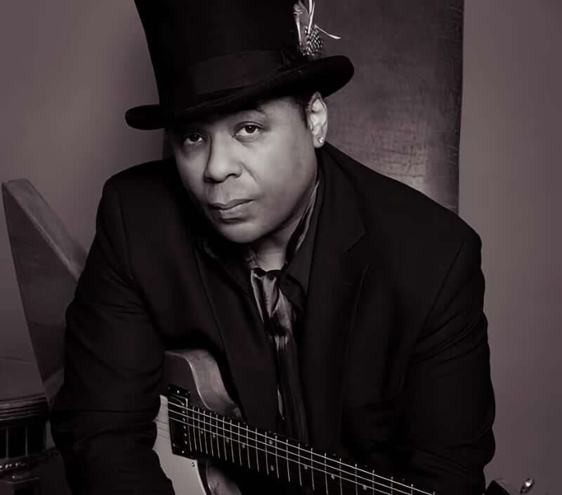 A black and white studio portrait of a medium-skinned African American man holding a Gibson Explorer guitar. He is wearing a black tophat with a feather in the hatband and a black suit jacket with a dark-colored satin tie.