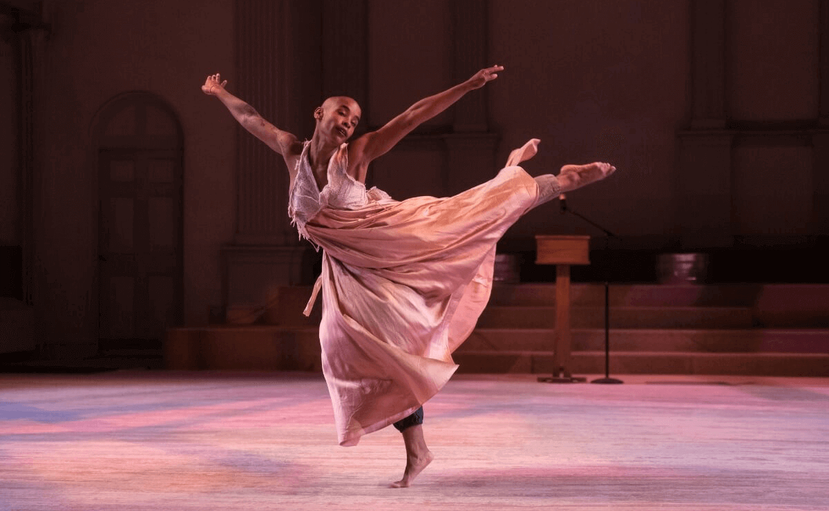 A dancer performs gracefully in a softly lit space with warm tones. The dancer, Jasmine Hearn, is a Black woman with a shaved head and bare feet, and she is wearing a flowing pink and white dress with subtle shades of purple. She is photographed mid-motion with one leg extended behind her and arms outstretched. Her expression conveys focus and emotion. The background features architectural elements, including columns and a wooden podium, suggesting a performance in a formal or historic venue. The floor is illuminated with subtle pink and purple lighting.