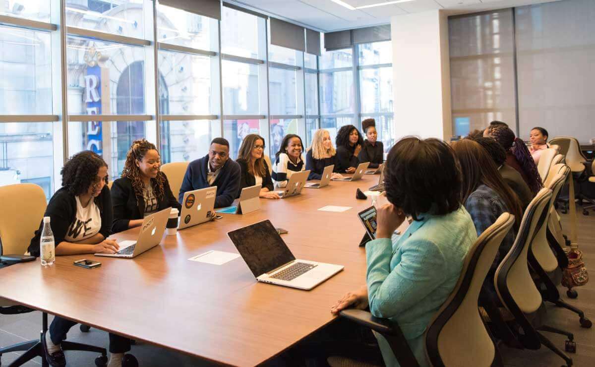 A group of over a dozen BIPOC leaders sit facing each other at a long conference table in a sunny boardroom. They each have a laptop open before them, and they are smiling and talking amongst themselves.