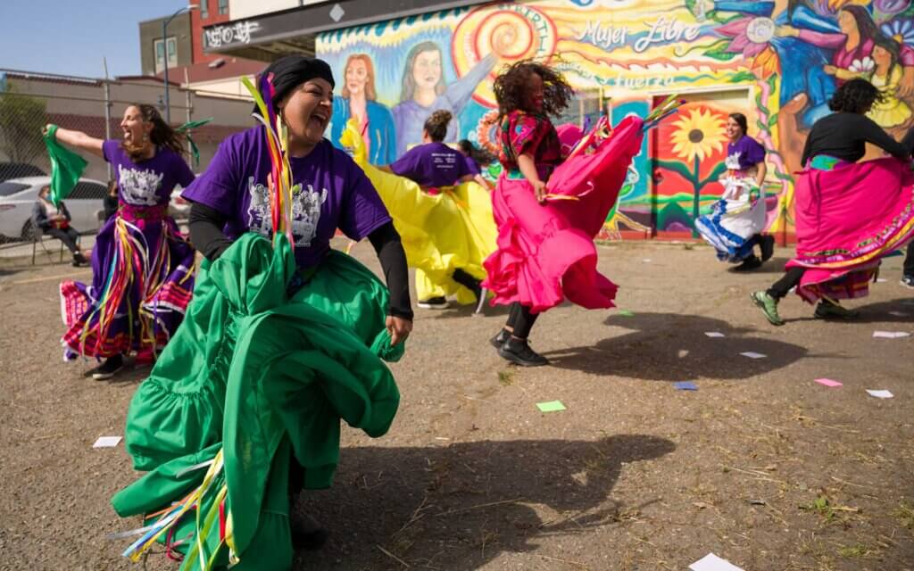 A woman in a purple top and green folkloric skirt laughs as she dances and moves the skirt and colorful ribbons. Three other women in colorful skirts (bright pink, yellow, and purple) are dancing in the background against a brightly colored mural with a sun and flowers. They are outdoors on a sunny day, and shadows of their bodies are cast upon the ground.