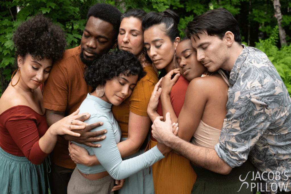 Seven dancers of different races embracing one another in a collective hug with their eyes softly closed.