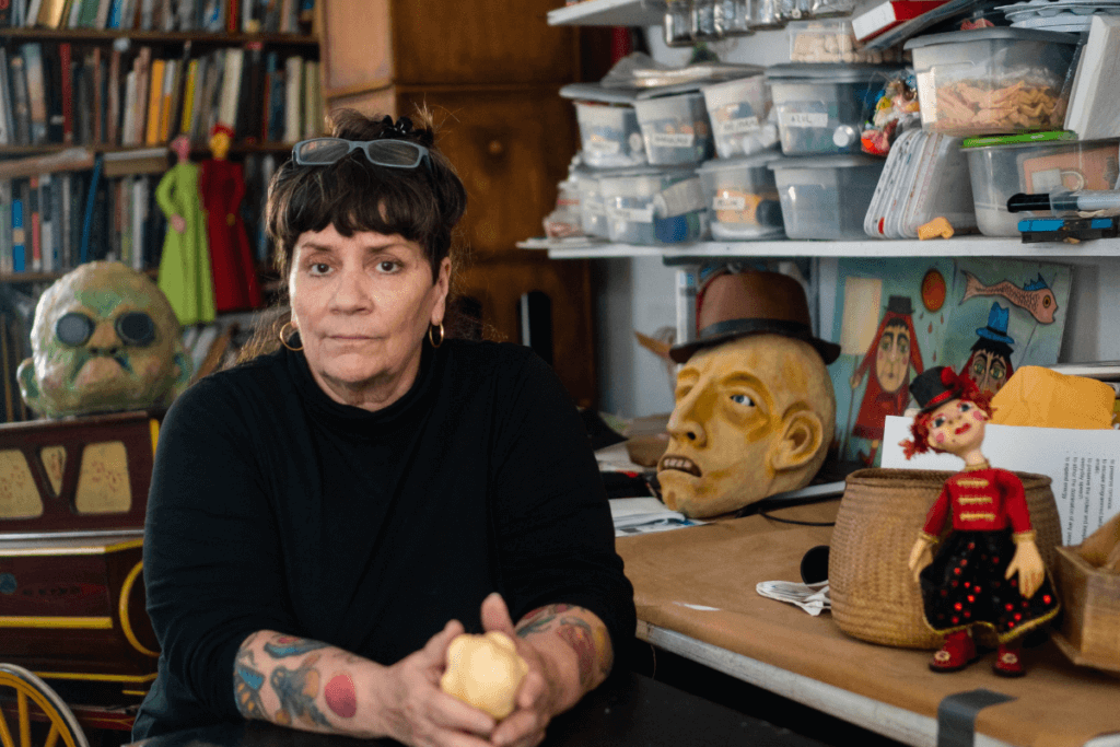 A light skinned mature woman dressed in black is seated looking straight at the camera. Behind her are books and masks and materials for creating puppets. In her hand she holds a small puppet head.