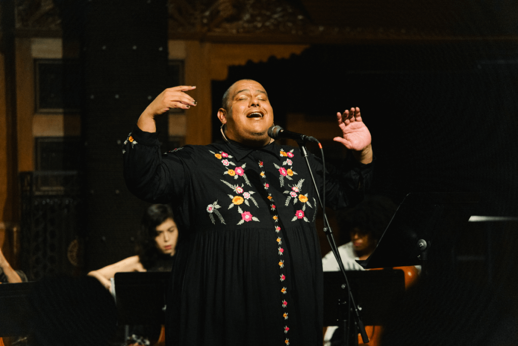 A medium-skinned trans femme person with her mouth open in front of a microphone. Her arms and hands are raised at face level. She is wearing a black robe-like dress with embroidered flowers of varying colors.