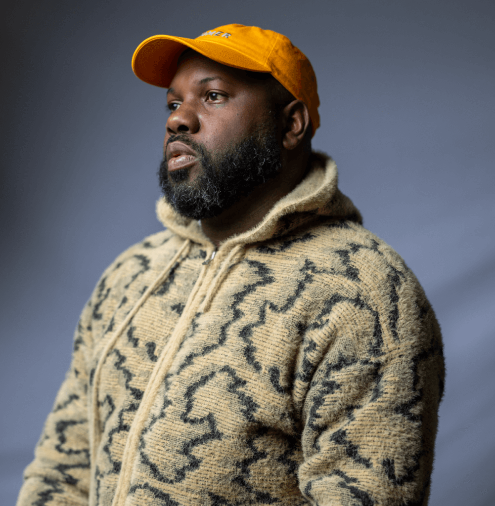 A dark-skin Black man, in a black and tan hoodie and an orange cap. It is a 2024 self-portrait of L. Kasimu Harris, a writer and artist.