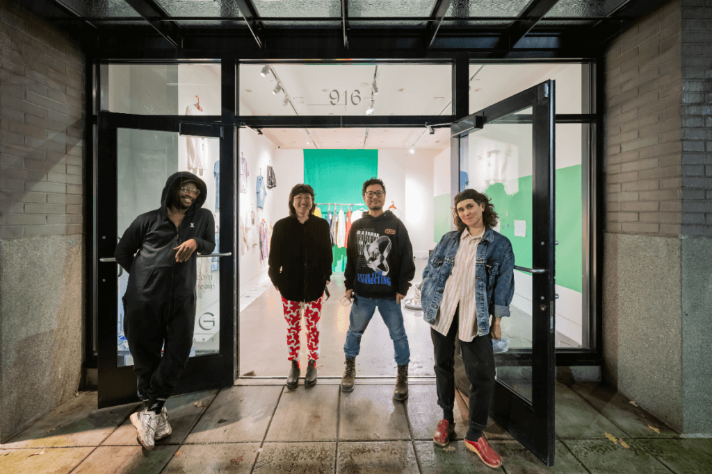 A group of four people outside a gallery with large windows. From left to right: a Black person in a black jumpsuit, an Asian person in red and white pants and black blouse, an Asian person in jeans and graphic hoodie, and a white person in black jeans, pink and white striped blouse and denim jacket.