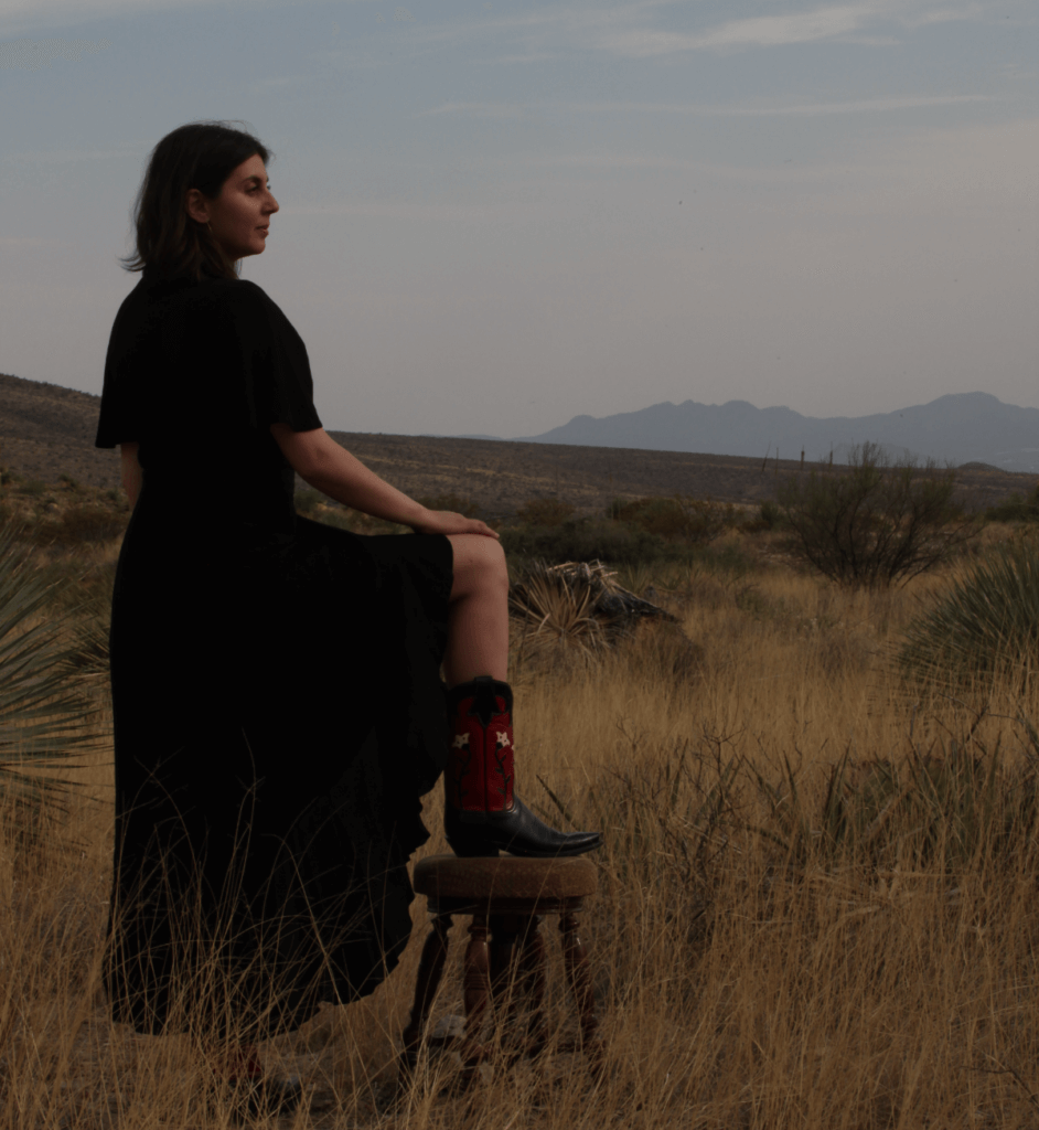 A woman in a black flowing dress and cowboy boots facing sideways, in front of a desert landscape. The wind is blowing her dress slightly, blending it into the desert shrubs at her feet.