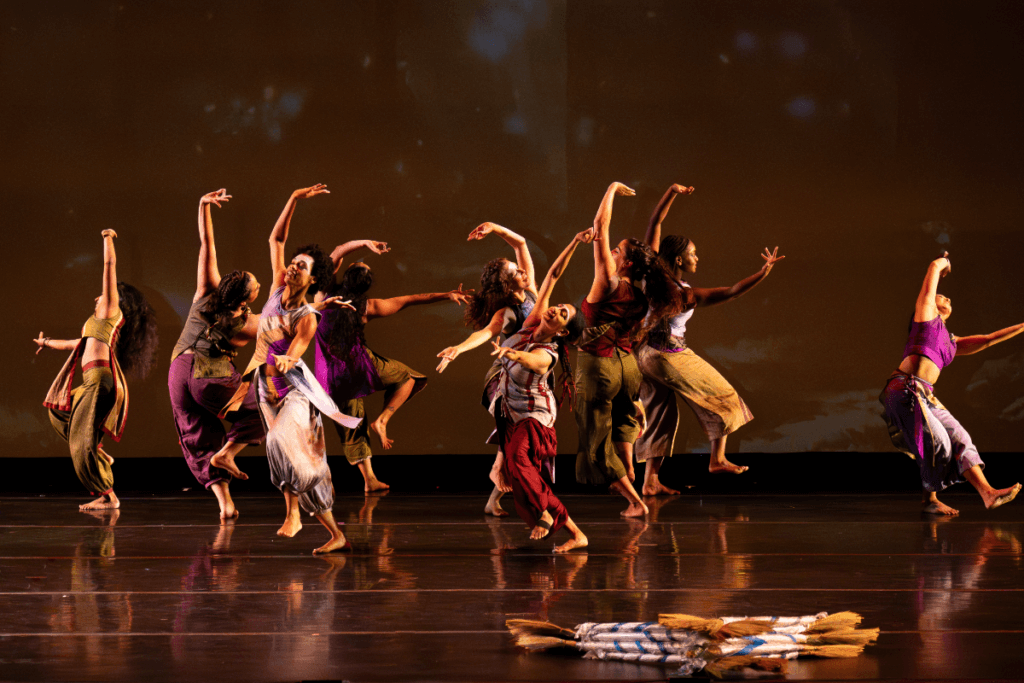 Nine femme dancers curve their arms in luscious extension, facing all directions but connected to each other in warmth. They are caught, in this photo, mid-movement, one knee bent and foot flexed, ready to take the next grounded step. Their skin is of varying brown skin tones with varying hair lengths and textures; their wear silk dupioni costumes (sleeveless tops and pants) in various colorblock shades of fuscia, lime green, lavender, silver, red, and amber. In the foreground on the right, there is a neat stack of short, ceremonial brooms, with silver and blue silk-wrapped handles. The background is a warm brown with soft small "fireflies" of light scattered throughout.