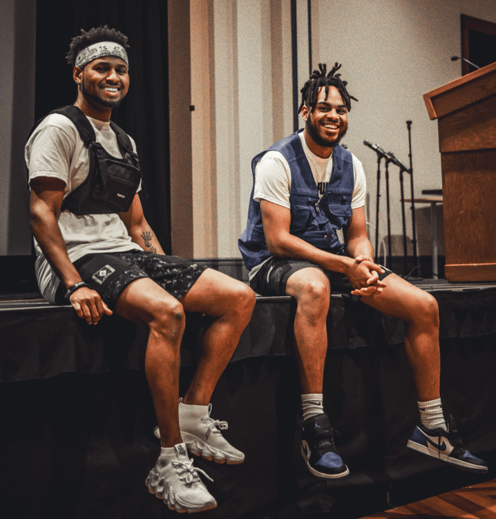 In this Image is two African American Men sitting on a stage after a live performance. On the Left hand side is Marcell Johnson With afro and white bandana, White t shirt with black vest and black shorts and white sneakers Host and Mc of the Yes Foundation Violence Prevention Symposium , on the right hand Side is Yusef Shelton with white t shirt, blue vest, black shorts, blue, black and white nike sneakers, who just Mc'd and performed music at Yes foundation Symposium where the pair end up being on local news station for their participation and involvement in bringing culturally relevant educational, fun and engaging energy to event held at Pitt University in collaboration with Pennsylvania number one violence prevention organization. Shot by Reece Miller.
