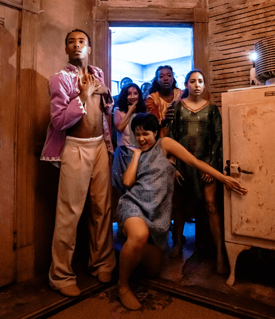 Five dancers pour out of a doorway towards us. To the far left, a medium-dark skinned dancer with short black hair stands with his hands over his chest. He is wearing a pink-lavender open shirt with tan pants. To his right is a medium-light skinned dancer wearing a white, sleeveless shirt and shoulder-length black hair. She is smiling with one hand on their chin. To the right of them stands a dark-skinned dancer with her mouth open in shock. She is wearing a cream and orange top and has her hand stretched out on the arm of a dancer standing in front of her who is wearing a green dress. The dancer in the green dress has a pensive expression. Centered, and in front, is a medium-skinned dancer wearing a blue-grey dress. They have one knee on the ground, the palm of their hand on their chin, and a joyful, mischievous expression on their face. They have their other arm extended grabbing onto a cabinet to their right.