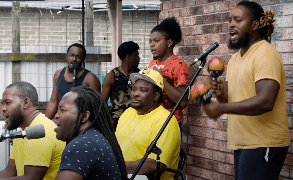 Seven dark skinned men perform music in the back yard of a brick house. They play drums and shakers, and some sing into microphones.