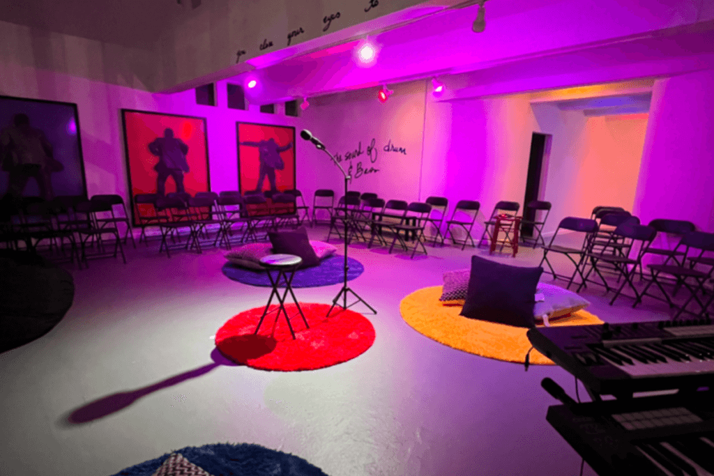 A lone microphone stand is spotlit is in the center of a small room lit with multi-colored lights. Pillows on the floor, colorful carpets and chairs surround the microphone stand.