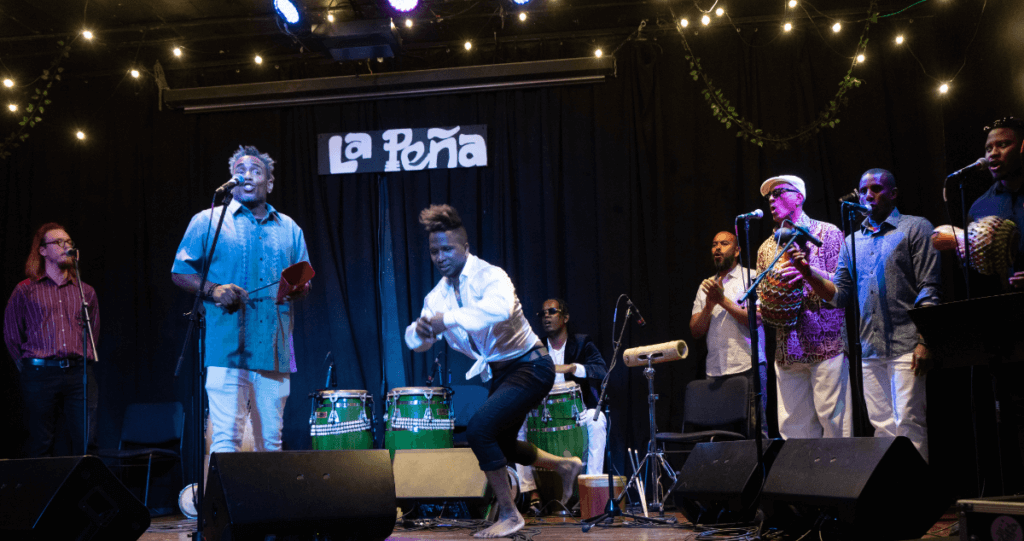 A group of musicians singing and playing instruments on a stage at La Pena Cultural Center. An Afro-Cuban male drummer playing congas. A dancer with arms lifted midway and his feet lifted off the ground. The lead singer, a medium-dark-skinned Cuban man, wears a grey Guayabera and is mid-song, facing the audience and microphone. Two musicians are playing large chekeres.