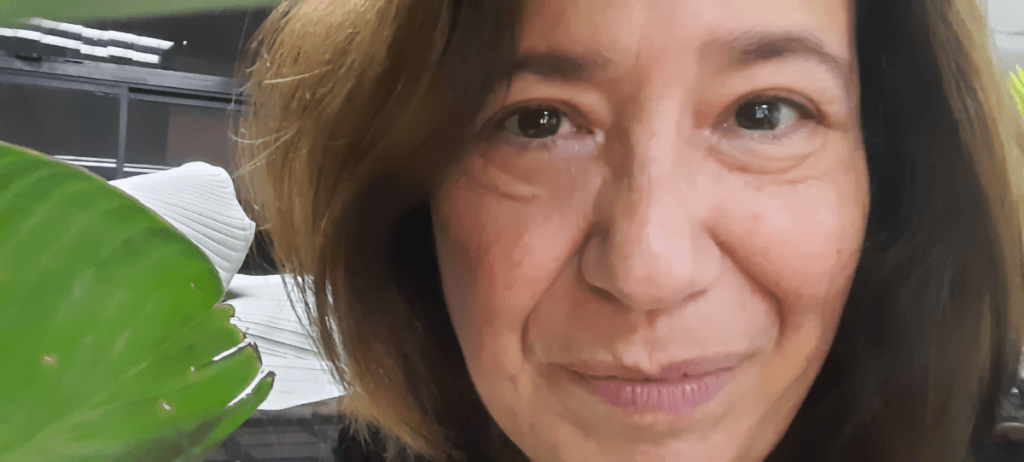 Selfie of woman with light brown face close up, medium brown hair, looking straight into the camera, slightly smiling with mouth closed, green large tropical leaves in the background.
