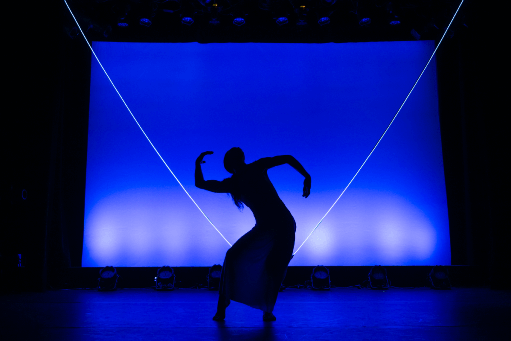 A dancing silhouette in a skirt with arched in an S curve spine and arms against a blue backdrop with a glowing “V” upstage.