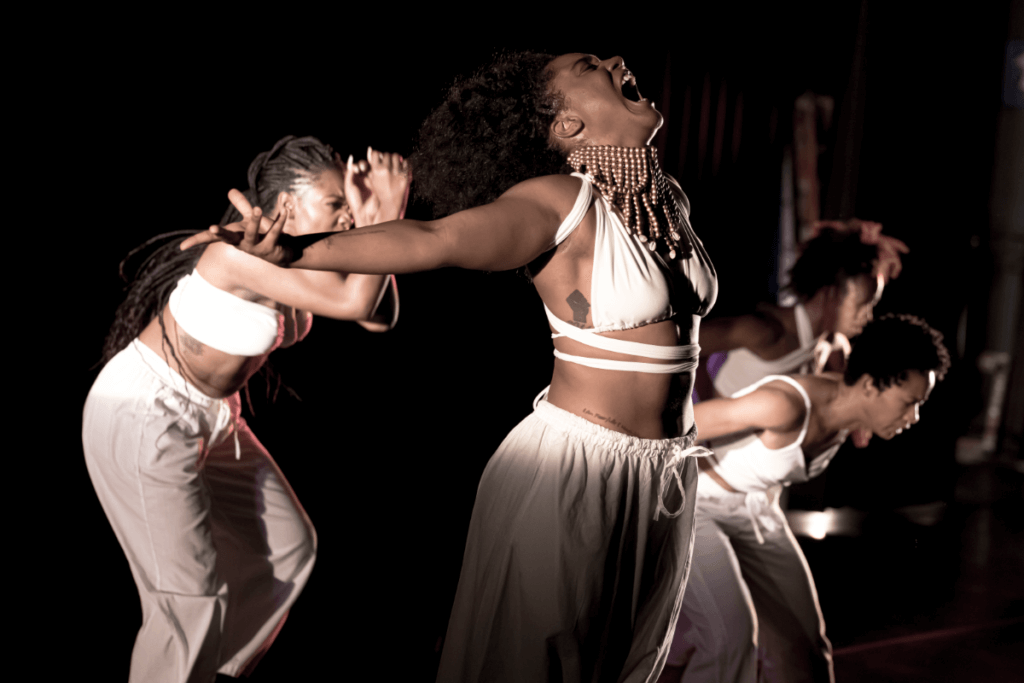 This image consists of four dancers. Chanice Holmes, a medium brown skinned African American female dancer wearing a white wrap crop top, white flowy pants and gold beaded choker with cowrie shells, stands facing to the side with both arms reaching out to the sides, head tilted back and mouth open in a mid-scream. Millenique Brown, a medium brown skinned African American female dancer wearing a white wrap crop top and white pants, stands facing to the side with arms bent in mid-reach up, torso bent forward and legs in mid-step. Catherine Caldwell, a medium brown skinned African American female dancer wearing a white crop tank and white flared pants, stands facing to the side with arms reaching side, torso bent forward and legs in mid-step. Jasmin Simmons-Edmond, a medium brown skinned African American female dancer wearing white wrap crop halter and white flowy pants, stands facing to the side with arms reaching side, torso bent forward and legs in mid-step.