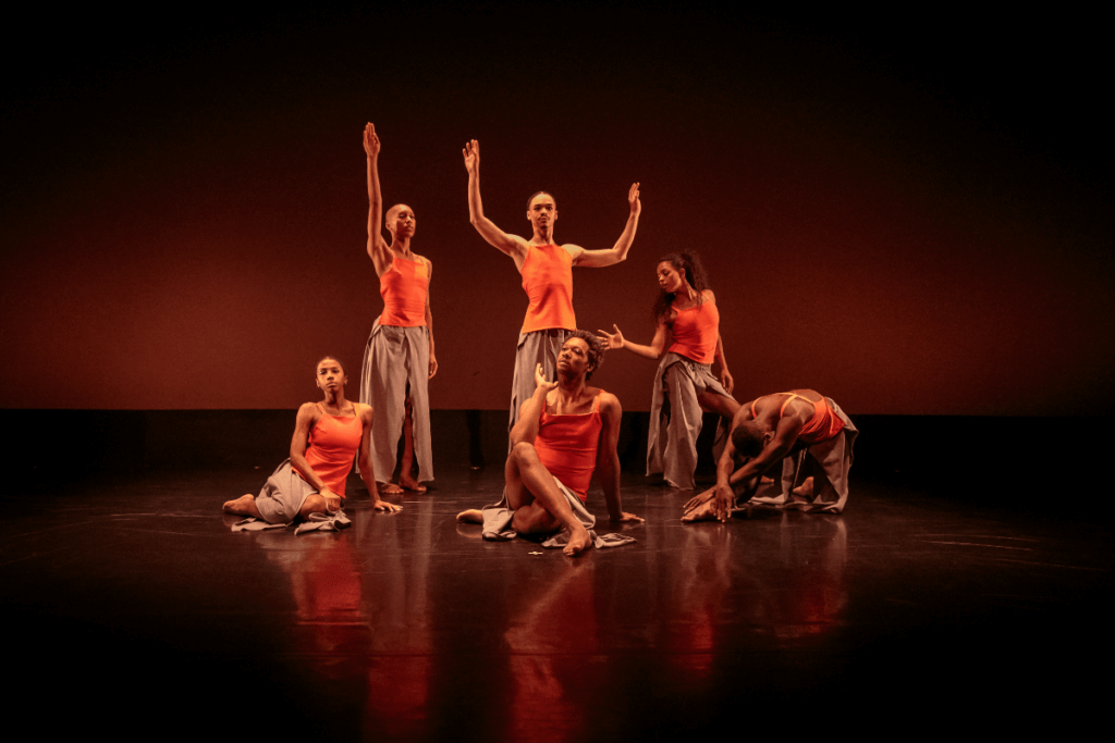 Six Black and Brown of mixed skin tones pose in a group tableaux. They are wearing uniform orange tank tops and grey pants with skirt panels draping below. They are on a stage with a black floor and cyc with faint orange light.