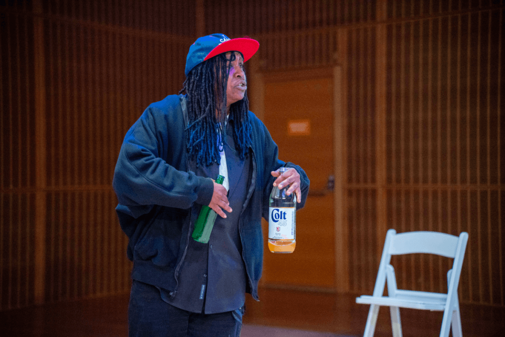 A dark-skinned man with braided dreads, wearing a dark jacket, a black shirt, and black pants. He also has on a two-tone baseball cap—blue with a red brim. In his left hand, he holds a large glass bottle of Colt 45 malt liquor, and in his right hand, a green glass bottle . Their facial expression is intense and engaged, as if they are mid-speech.