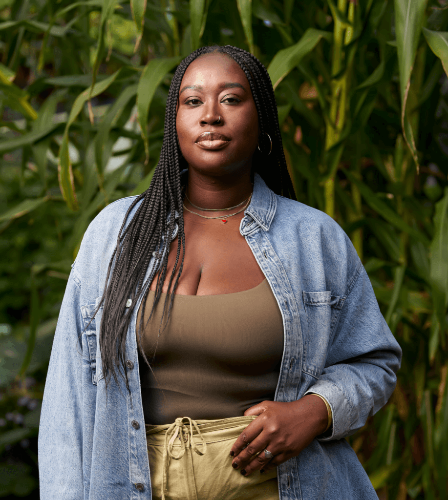 Nana Kumi, a dark-skinned Black woman, stands in a lush community garden on a crisp fall morning, with a strong, rested expression. She wears a green bodysuit and green linen pants, symbolizing her connection to the earth, and a denim shirt honoring the legacy of Black laborers. Her box braids fall over her right shoulder, while the left side is tucked behind her back. One hand rests on her hip, thumb gently tucked into her waistband. She stands grounded and centered in the community garden of a beloved elder, surrounded by plants and the quiet power of ancestral presence.