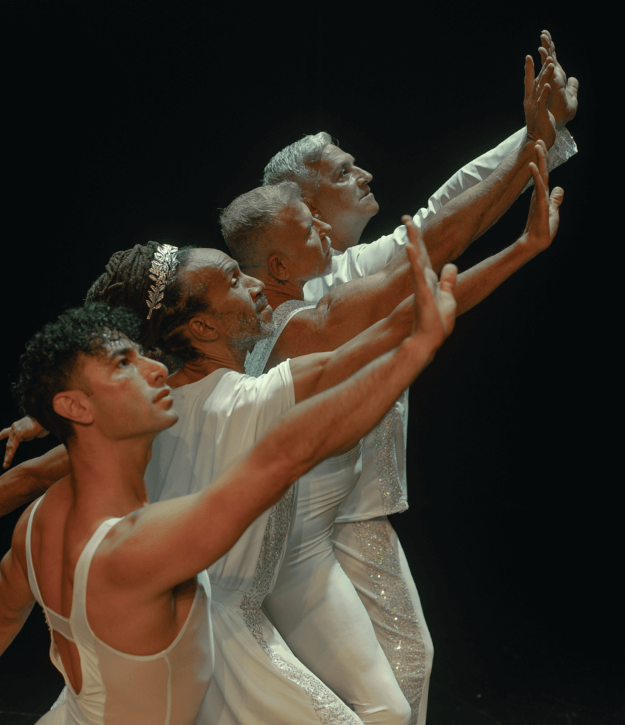 The image shows four dancers in a unified, graceful pose, their arms extended forward in a reaching gesture with flexed hands. Each dressed in various styles of shimmering white costumes, the dancers’ bodies are bathed in soft light that highlights their forms. The front dancer, younger with curly brown hair, gazes upward, while the three elder dancers behind echo the same gesture, their expressions full of intensity and warmth. The dark backdrop amplifies the sense of unity and reverence in this intergenerational moment, evoking themes of strength and connection.