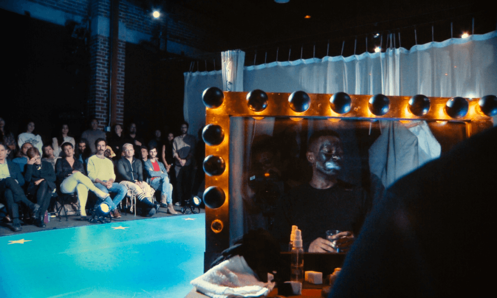 In the foreground on the right, is a large, illuminated dusty dressing room mirror outlined with round black-and-gold bulbs. Reflected in the mirror is a Black male performer, seated wearing a black shirt. His face is painted in black makeup—with exaggerated white paint around the mouth and eyes. He holds a glass in one hand, and his expression appears still and solemn. In front of him on the dressing table, there are a white rag with a red makeup stain, a satin black glove, a clear spray bottle and a sponge soaked with black makeup. Behind him, the mirror reflects a hanging white curtain and white dress shirt and grey jacket hanging from a hook. To the left, in the background, sits an entirely light- skinned audience in a dimly lit space. The crowd is mixed in gender and dress, with people sitting closely in folding chairs. They face the stage, looking towards the performer. Their expressions range from neutral to serious. The walls behind them are dark with exposed red brick pillars, and overhead lights cast a cool glow on the bright blue stage floor, which is outlined with a few white stars.