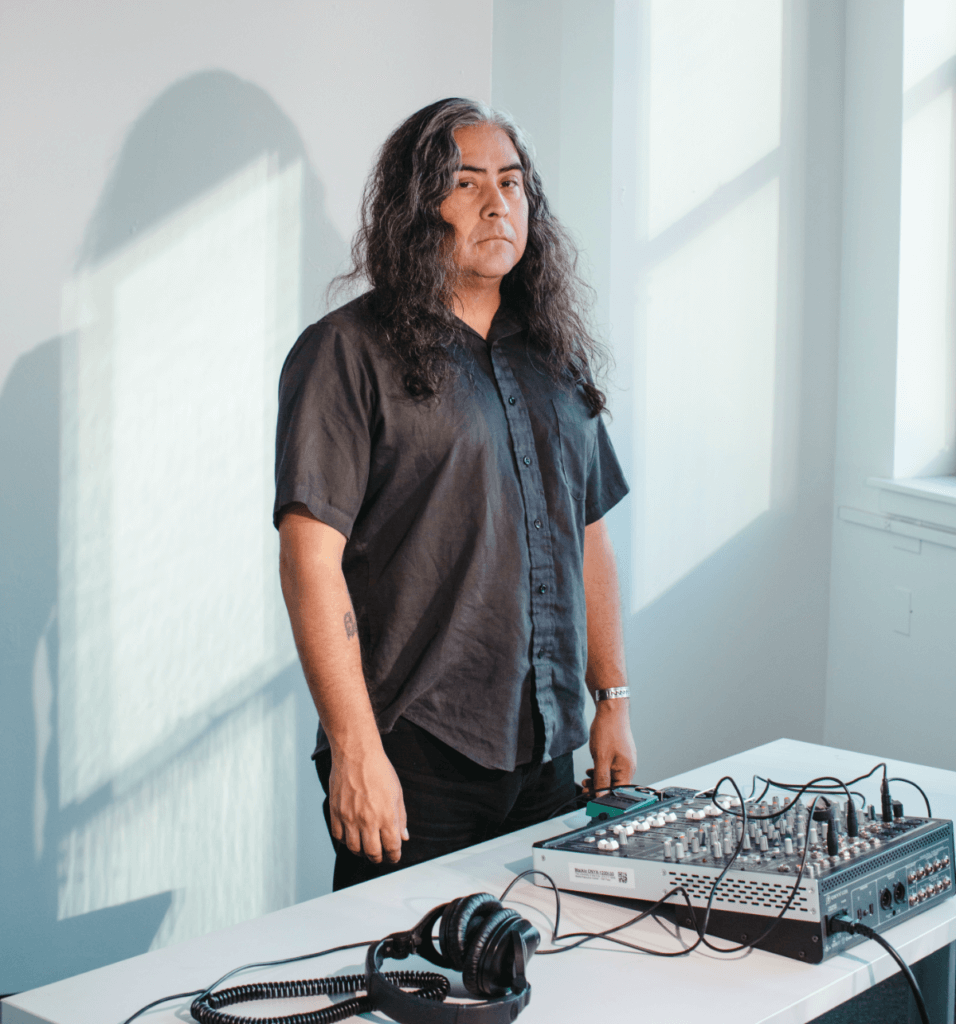 Composer Raven Chacon stands upright in the center of a well-lit room, wearing a black short-sleeved button down and black pants. He is standing behind a folding table on which sits an audio mixing console and a pair of black headphones. He is staring into the camera, with light from the window framing his figure on the wall behind him.