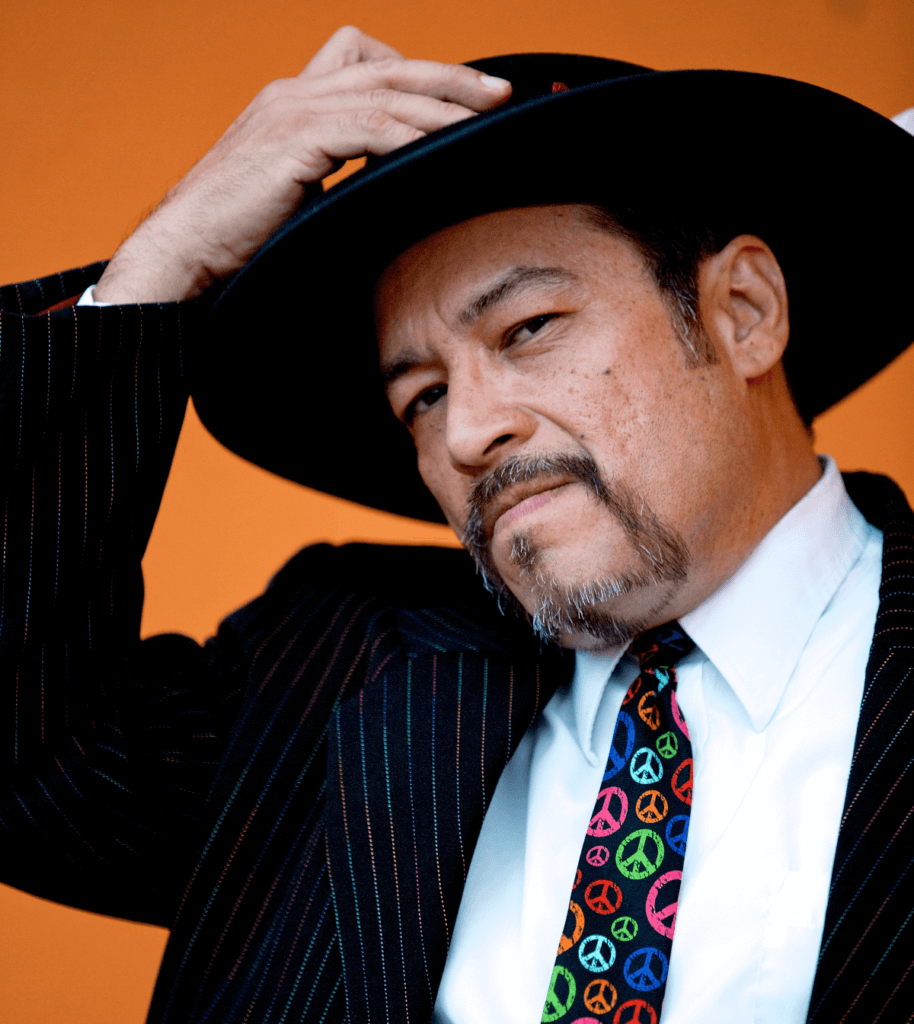 A medium skinned Chicano man dressed in a pin-striped suit, with a white shirt and a multi-colored tie that features round peace symbols. His facial features include a moustache that connects to a goatee. His left hand holds his left lapel, as his right hand reaches for his black, wide-brimmed pork pie Tando (hat), as if about to remove it from his head. The background is covered in a brown/orange tone.
