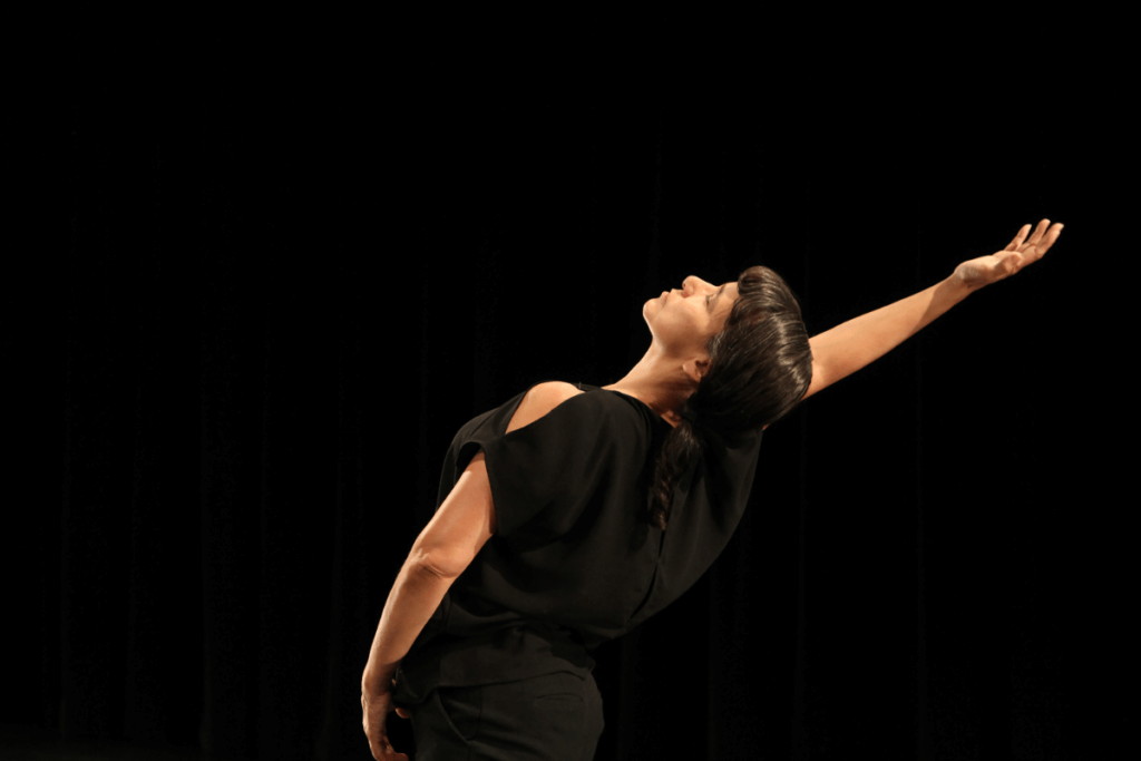 Against a jet black background a Native woman with medium brown skin, dark brown straight hair in a single loose braid and regal facial expression arches back in a dancerly position with her right arm raised high, looking up. She wears a black outfit with a sleeveless top.