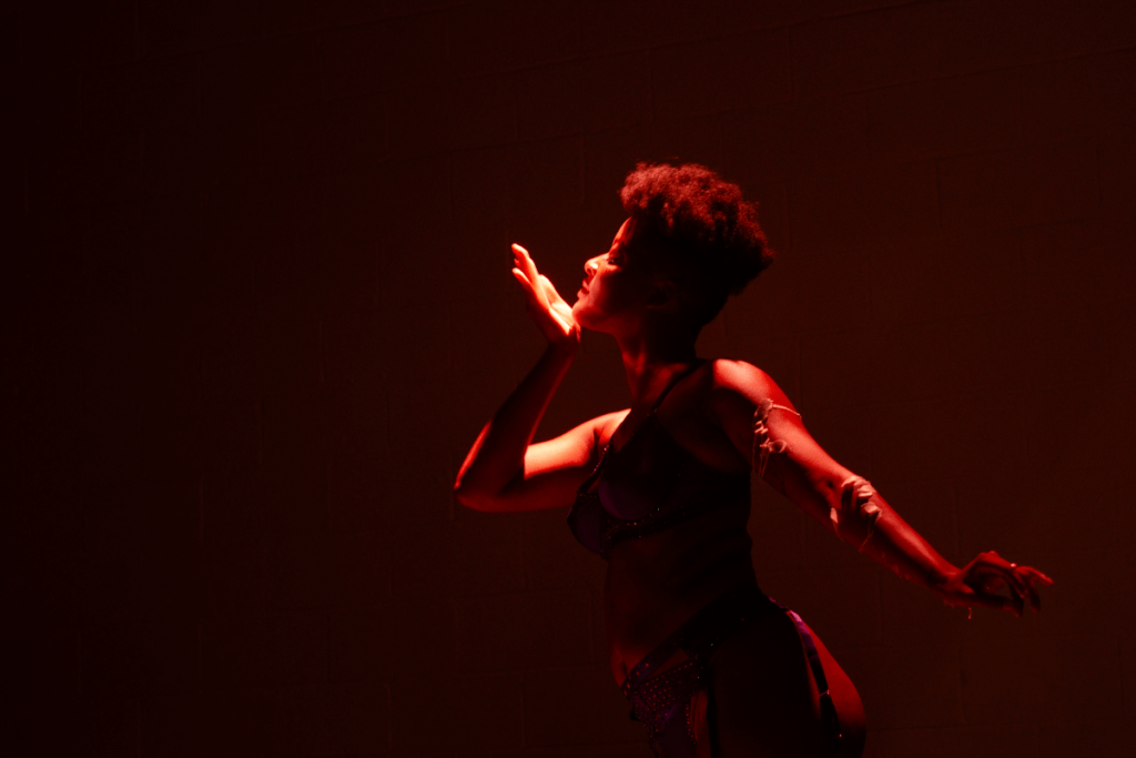 A brown-skinned woman mid-movement dancing. Her right arm is bent at the elbow, heel of palm cupping her chin as she looks to the right. Her left arm is outstretched, and white wax can be seen clinging to her skin on that arm. She is wearing purple rhinestoned bra, garters, and thong, which are shadowed in the spotlight.
