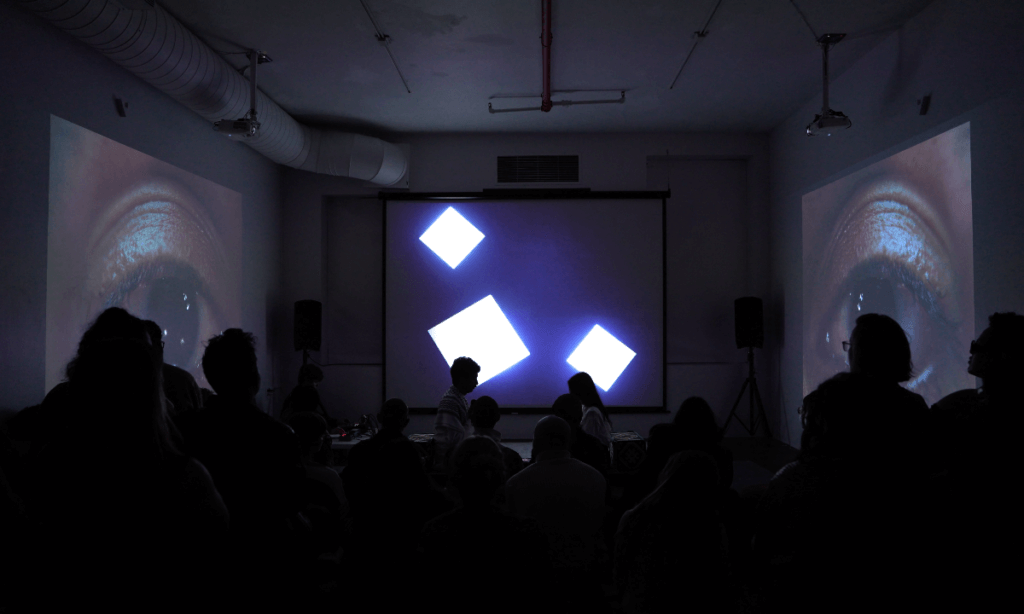 A dimly lit room is filled with an audience seated on the floor, facing three large projections that span the front and side walls. The central projection shows animated white squares of varying sizes floating against a black background. On the left and right walls, close-up videos of a brown-skinned, South Asian person's eye dominate the space, each with a glistening reflection of stars or lights in the pupil. In front of the central screen, silhouetted audience members are visible, with a few standing or moving about. Among them, one person with medium-dark skin and short curly hair appears to be operating audiovisual equipment on a table. Two ceiling-mounted projectors and large speakers are visible above and around the room. The atmosphere illuminates the activation of an immersive audiovisual performance and video installation.