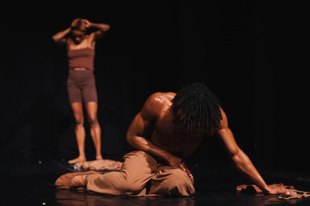 Two brown-skinned dancers are pictured against a black backdrop. The dancer on the far left background stands looking down, dressed in a close-fitting brown tank top, brown biker shorts, and flesh-tone ballet shoes. The dancer on the right in the foreground sits on the floor supported by their left arm, looking down, face covered by short hair, bare-chested, wearing brown pants and flesh-tone ballet slippers.